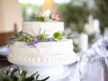 Dwupoziomowy tort weselny ozdobiony kwiatami, ziołami i oliwkami. Delikatne kremowe wykończenie i naturalne dekoracje podkreślają wyjątkowe smaki tortów weselnych.
