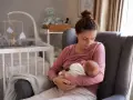 Matka karmi piersią noworodka w fotelu. W tle widać łóżeczko dziecięce. To piękny obrazek z okresu po porodzie.