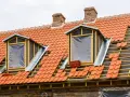 Trwa wymiana okna dachowego. Dach pokryty czerwona dachówką, widoczne drewniane elementy konstrukcji i nowe okna z folią.