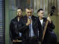 Rock n roll band in Lederjacken. Sänger hält Mikrofon, Bassist spielt Kontrabass, Schlagzeuger hält Sticks.