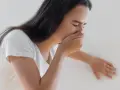 Kobieta w białej koszulce zakrywa usta dłonią, odczuwając mdłości. To częsty objaw, kiedy mdłości w ciąży dają o sobie znać.