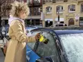 Woman in tan coat uses yellow electric ice scraper on frosted car window.