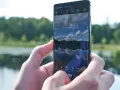 Dłonie trzymają smartfon Samsung S10, na ekranie którego widać odbicie nieba i lasu w spokojnej wodzie.