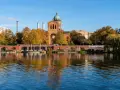 Herbstliche Bäume spiegeln sich im Wasser des Engelbeckens Berlin. Ein Café mit Außenbereich und eine Kirche mit Kuppel sind zu sehen.