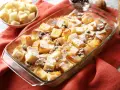 Pudding chlebowy w naczyniu, posypany cukrem pudrem i rodzynkami, obok miseczka z pokrojonym chlebem.