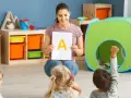 Nauczycielka pokazuje literę "A" dzieciom, stosując metodykę wprowadzania litery w przedszkolu. Jedno z dzieci podnosi rękę.