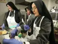 Nuns in a kitchen preparing food. One nun smiles while serving, another holds a fork, and a third looks on. This scene might answer the question: do nuns eat meat?