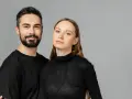 Para, która wygląda jakby mogła nosić ubrania unisex. Mężczyzna z brodą i kobieta z długimi włosami, oboje w czarnych, prostych ubraniach.