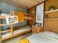 Wnętrze chałupy kempingowej z łóżkiem piętrowym i pojedynczym, żółtym kocem i torbą z napisem "MAKING PEOPLE SURF".
