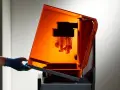 A gloved hand opens the orange lid of a stereolithography 3D printing machine, revealing intricate parts being built layer by layer.