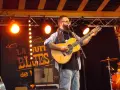 Un homme joue de la guitare sur scène, éclairé par des projecteurs. Le panneau derrière lui annonce "La Nuit du Blues", évoquant la naissance du blues.