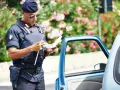 Agente di polizia italiana con giubbotto antiproiettile e berretto blu, controlla un verbale vicino a un'auto blu. Potrebbe essere un controllo legato all'art. 180 cds.