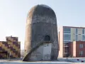 Der Trudelturm Berlin, ein futuristisches Betonbauwerk, steht neben modernen Gebäuden. Eine Außentreppe führt zu einer runden Tür.