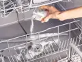A hand pours white powder from a jar into an open dishwasher, demonstrating how to clean a dishwasher naturally.