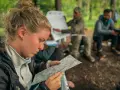 Frau studiert Karte, während Männer im Hintergrund bei einer Ausbildung im Wald sitzen. Solche intensiven, kurzen **ausbildungen die nur 1 jahr dauern** sind ideal für schnelle Karriereschritte.
