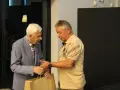 Zdzisław Kuźniar syn, starszy pan w niebieskiej marynarce, wręcza prezent młodszemu mężczyźnie w beżowej koszulce.