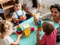Zwei Frauen und drei Kleinkinder spielen mit bunten Spielsachen. Eine Frau hilft einem Kind, Türme zu stapeln, was eine tolle Methode ist, um Kinder zweisprachig zu erziehen.