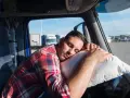 Ein LKW-Fahrer schläft auf einem Kissen im Führerhaus. Das Bild thematisiert die Einhaltung des Arbeitszeitgesetzes für LKW-Fahrer.