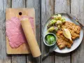 Förberedelser för schnitzel recept: tunt bankat kött på skärbräda, bredvid en tallrik med schnitzel, potatissallad och gurka.