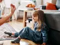 Dziewczynka w dżinsowym ogrodniczku siedzi na podłodze, uśmiecha się. Mama wyciąga rękę. Brak oznak agresji u dzieci.