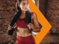 Donna sudata in abbigliamento sportivo rosso, con un asciugamano nero, si riposa dopo l'allenamento. Con il caldo i battiti aumentano, ma lei è determinata.