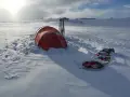 Ein rotes Wintercamping Zelt steht im Schnee, daneben Skier und ein Schlitten. Die Landschaft ist weit und verschneit.