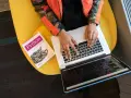 Osoba w pomarańczowej kurtce pisze na laptopie obok książki o Pythonie, siedząc na żółtym krześle.