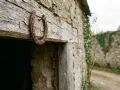 Un vieux fer à cheval pointes en bas, rouillé, est cloué sur une poutre en bois patiné, devant une bâtisse en pierre recouverte de lierre.