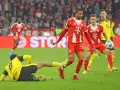 Intensywna akcja na boisku podczas meczu składy: Borussia Dortmund – Bayern Monachium. Piłkarz Bayernu w czerwonej koszulce próbuje odebrać piłkę leżącemu zawodnikowi BVB.