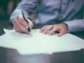 Ręka pisze długopisem na papierze, przygotowując dokumenty. Dowiedz się, jak założyć sprawę w sądzie, wypełniając niezbędne formularze.