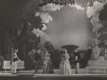 Scena z przedstawienia teatralnego, gdzie aktorzy w strojach z epoki odgrywają scenę w ogrodzie. Reżyser teatralny Szyfman zadbał o szczegóły scenografii.