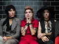 Trzech członków Palaye Royale w stylowych, rockowych strojach siedzi obok siebie na tle czarnych płytek.