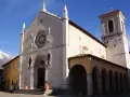 A grand church with a rose window and bell tower stands in a town square, with snow-capped mountains in the distance. This is Nursia, a historic Italian town.