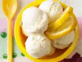 Erfrischendes Buttermilch Zitronen Eis in einer gelben Schale, garniert mit Zitronenscheiben. Ein gelber Löffel liegt daneben.