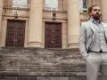 Elegancki mężczyzna w jasnej, kratkowanej marynarce i kamizelce, z chusteczką w kolorze śliwkowym w kieszeni, stoi na schodach przed budynkiem.