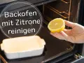 Backofen reinigen mit Zitrone: Eine Hand hält eine halbe Zitrone über eine leere Auflaufform im dunklen Backofen.
