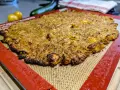 Une galette complète au four, dorée et croustillante, sur un tapis de cuisson en silicone. Des tomates jaunes et une courgette sont floues en arrière-plan.