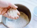 Mano sosteniendo arroz para calcular el tiempo cocción arroz perfecto. Olla con arroz y taza de agua al lado.