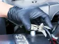 Gloved hand applying WD40 to a car battery terminal to prevent corrosion and ensure a good connection.