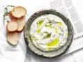 Kremowe **labneh co to**? Gęsty serek jogurtowy z oliwą, ziołami i pieczywem, podany na gazecie.