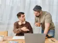 Ojciec rozmawia z synem, pokazując mu, jak zmotywować dziecko do nauki. Na stole laptop, zeszyt i kubek kawy.