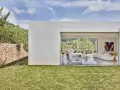 Exterior de casa ibicenca con césped verde y muro de piedra. Interior moderno con cocina y salón abiertos al paisaje.