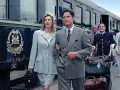 Elegancka para wychodzi z luksusowego pociągu, rozpoczynając swoje podróże koleją. W tle widać obsługę i innych pasażerów.