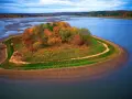 Jesienna wyspa na zalewie Brody, porośnięta kolorowymi drzewami i trawami, otoczona błękitną wodą.