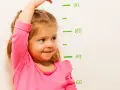 Uśmiechnięta dziewczynka w różowej bluzce mierzy wzrost przy tablicy z podziałką. Pytanie: co podawać dziecku na wzrost?