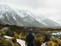 Une personne avec un sac à dos fait une randonnée sous la pluie, face à des montagnes enneigées. Le chemin en bois serpente à travers une végétation basse.