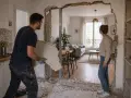 Un homme retire un morceau de plâtre, aidant à casser un mur non porteur. Une femme observe la transformation de l'espace.