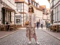 Eine Frau trägt eine leichte Jacke zum geblümten Kleid und weiße Sneaker. Sie steht auf einer Kopfsteinpflasterstraße vor Fachwerkhäusern.