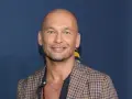 Tomasz Wygoda, łysy mężczyzna z niebieskimi oczami, uśmiecha się, ubrany w kratowaną marynarkę i naszyjnik.