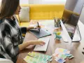 Młoda kobieta pracuje przy komputerze, korzystając z tabletu graficznego. Na biurku leżą próbki kolorów, ołówki i figurki. To idealne miejsce do rozwijania kreatywnych kierunków studiów.